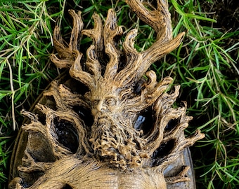 Puede incluir: Una escultura de resina marrón y dorada de un espíritu del árbol con cuernos, barba y un torso humanoide. La escultura está rodeada de hojas y flores.