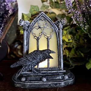 Könnte beinhalten: Ein schwarzer Rabe sitzt auf einem gotischen Fensterrahmen mit einer brennenden Kerze im Inneren. Das Fenster ist aus Milchglas und hat ein dekoratives Design. Der Rabe blickt mit offenem Schnabel zum Betrachter.