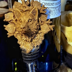 May include: A gold-toned bottle stopper featuring a detailed sculpted fox head surrounded by leaves. The stopper is attached to a silver-coloured metal rod.