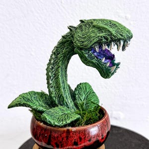 Op de afbeelding: Een groene, gestileerde plantensculptuur met een grote, open mond en scherpe tanden. De plant staat in een rood-bruine keramische pot met groene bladeren en mos. De sculptuur staat op een zwarte, ronde basis.