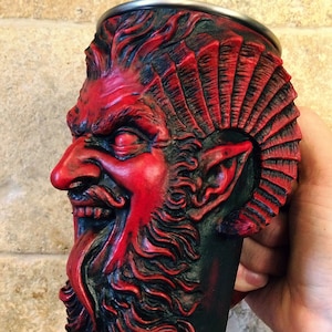 May include: A red and black ceramic mug shaped like a demon's head with a beard and horns. The mug has a detailed design and is perfect for serving beverages.