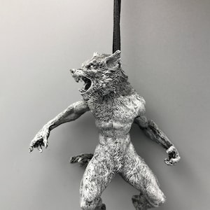 Könnte beinhalten: Eine silberne Werwolf-Figur, die an einem schwarzen Band hängt. Der Werwolf ist in einer knurrenden Pose dargestellt, mit offenem Maul und sichtbaren Zähnen. Die Figur ist detailliert mit Fell und Muskeldefinition.