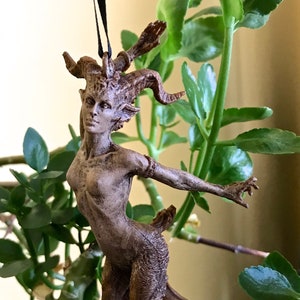 May include: A brown, detailed figurine of a female creature with horns, hooves, and a human-like torso. The figurine is hanging from a black cord.
