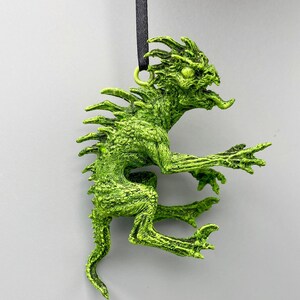 May include: A green, textured, plastic ornament of a mythical creature with a long tail and spiky back. The ornament is hanging from a black string.