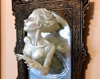 May include: A white, translucent, female figure appears to be emerging from a silver, ornate picture frame. The frame is dark brown with intricate carvings.