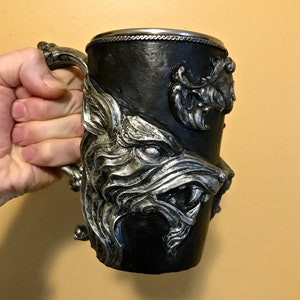 Puede incluir: Una taza de cerámica negra con un diseño de cabeza de lobo plateado. La taza tiene un borde plateado y un asa plateada.