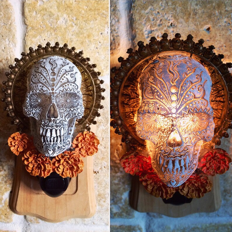 May include: A white and gold sugar skull nightlight with a floral design. The skull is surrounded by marigold flowers and is mounted on a wooden base.