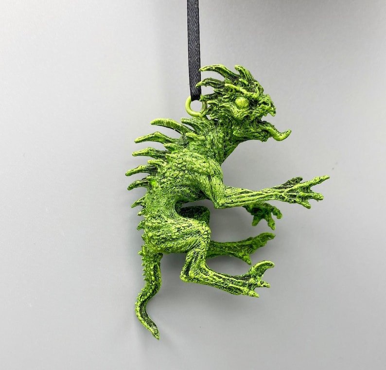 May include: A green, textured, plastic ornament of a mythical creature with a long tail and spiky back. The ornament is hanging from a black string.