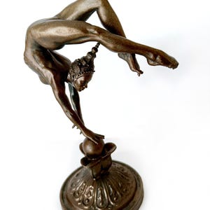 May include: A bronze sculpture of a woman in a contorted pose, balancing on a sphere. The woman is wearing a crown and has her arms outstretched. The sculpture is on a decorative base.