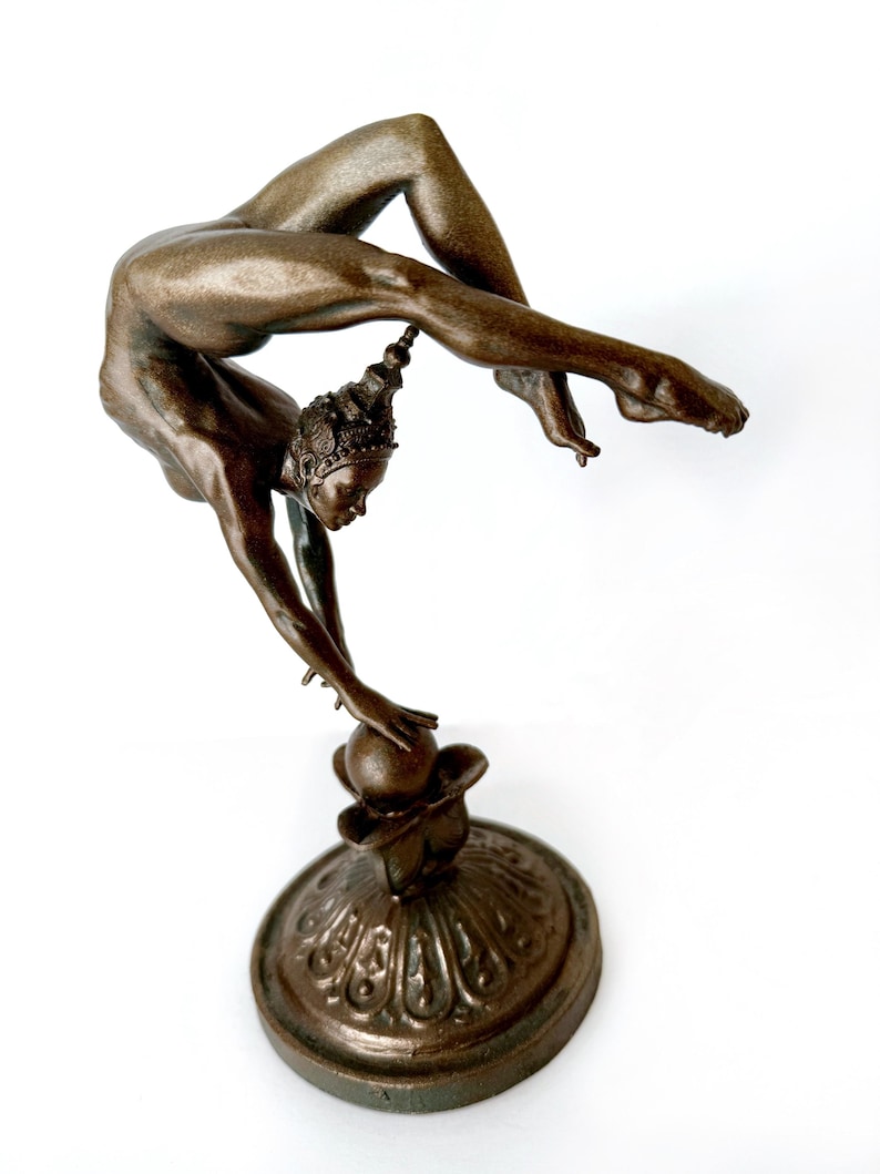May include: A bronze sculpture of a woman in a contorted pose, balancing on a sphere. The woman is wearing a crown and has her arms outstretched. The sculpture is on a decorative base.