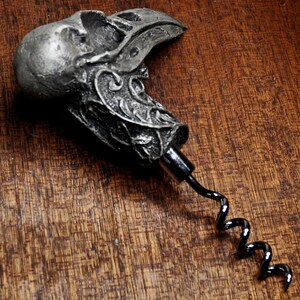 May include: A silver-colored metal corkscrew with a detailed skull and bird design. The corkscrew has a black metal spiral.