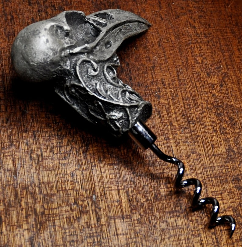 May include: A silver-colored metal corkscrew with a detailed skull and bird design. The corkscrew has a black metal spiral.