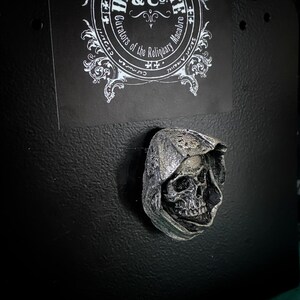 May include: A silver-colored skull wearing a hooded cloak, a gothic-style decorative item.