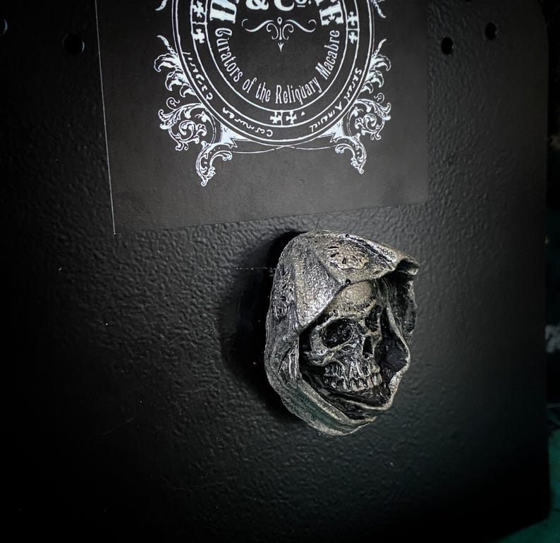 May include: A silver-colored skull wearing a hooded cloak, a gothic-style decorative item.