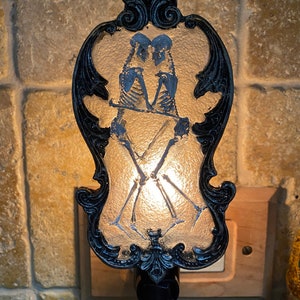 May include: A black and white nightlight with a decorative frame and a design of two skeletons holding hands. The nightlight is plugged into an outlet.