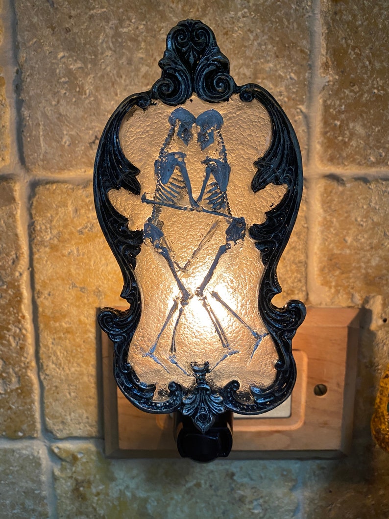 May include: A black and white nightlight with a decorative frame and a design of two skeletons holding hands. The nightlight is plugged into an outlet.