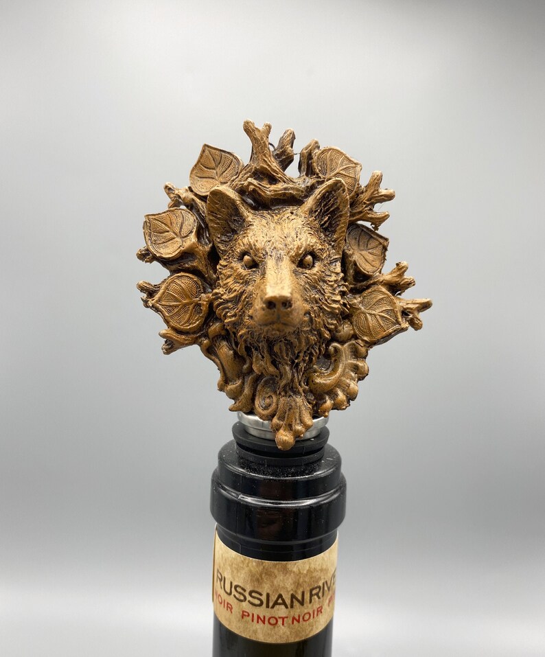 Op de afbeelding: Een gouden wijnflesstopper met een gedetailleerd wolfskopbeeld omringd door bladeren en takken.