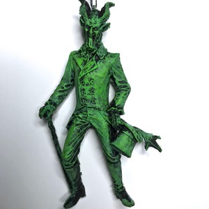 May include: Green, demonic-looking figure ornament with horns, a top hat, and a cane. The figure is wearing a suit and has a long, pointed tongue.