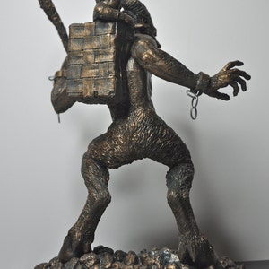 May include: A bronze sculpture of a goat-like creature carrying a small child on its back. The creature is chained at the wrist and has a basket on its back. The sculpture is set on a base of small rocks.