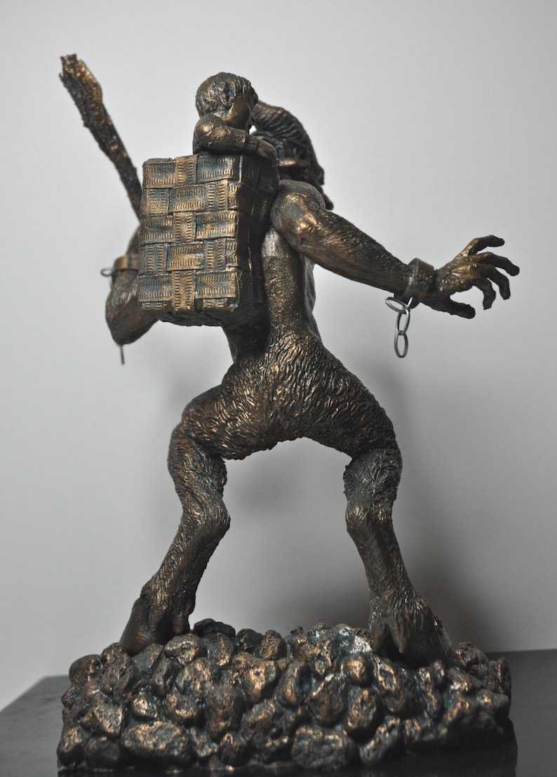 May include: A bronze sculpture of a goat-like creature carrying a small child on its back. The creature is chained at the wrist and has a basket on its back. The sculpture is set on a base of small rocks.