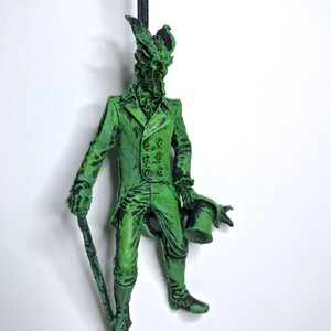 May include: A green figurine of a creature wearing a suit and holding a top hat and a cane. The creature has a long snout and horns.