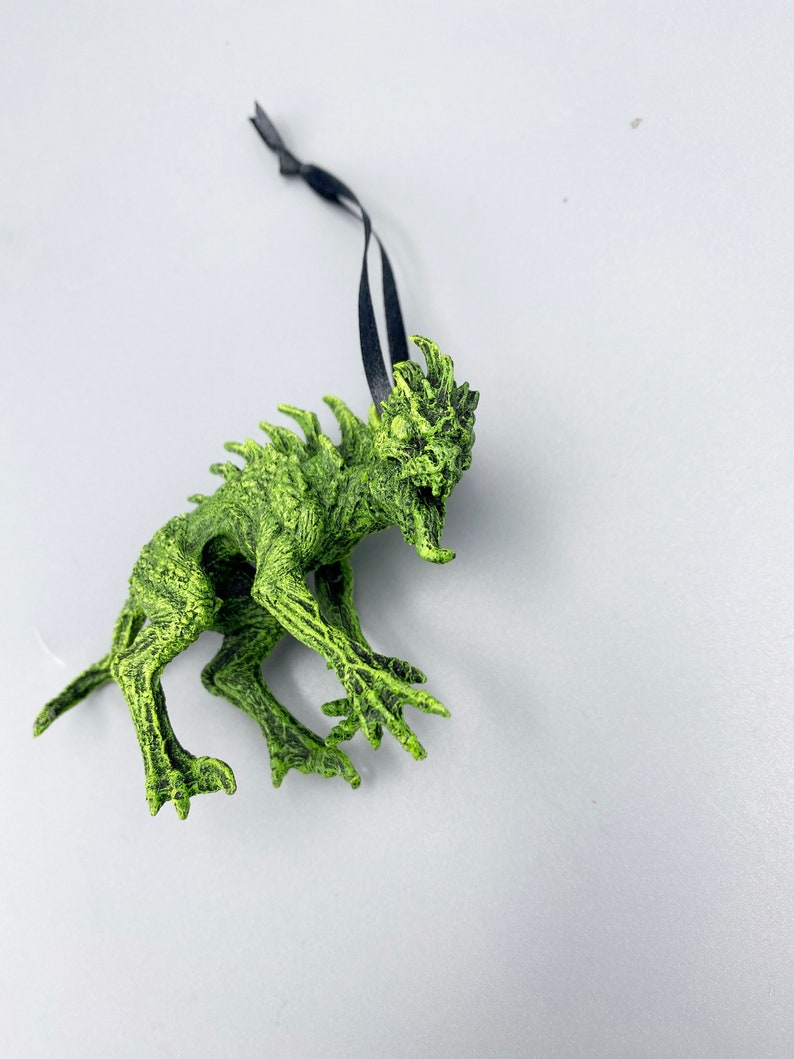 May include: Green monster ornament with a black ribbon for hanging. The monster has sharp teeth and claws.