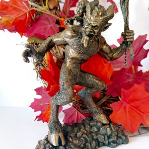 May include: A bronze statue of a horned, goat-like creature with a child on its back. The creature is holding a stick in its right hand and has a chain attached to its wrist. The statue is set against a background of red fall leaves.