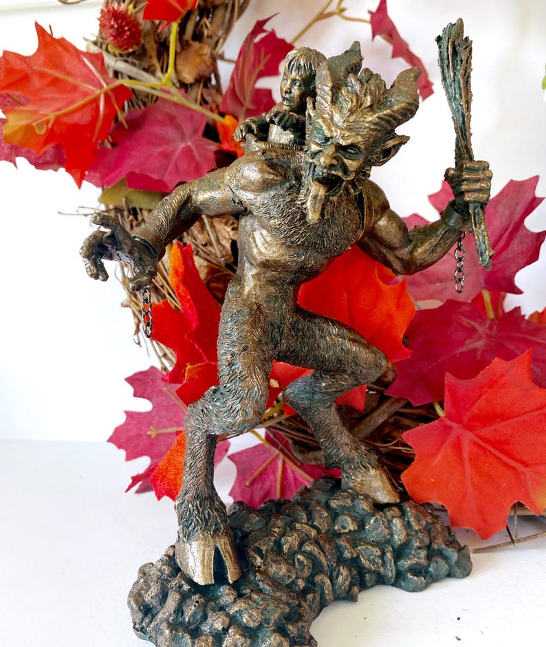 May include: A bronze statue of a horned, goat-like creature with a child on its back. The creature is holding a stick in its right hand and has a chain attached to its wrist. The statue is set against a background of red fall leaves.