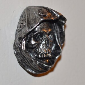 May include: A silver-colored skull with a hooded cloak, the skull has a detailed face with visible teeth and eye sockets.