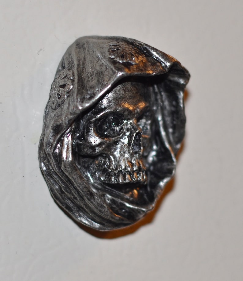 May include: A silver-colored skull with a hooded cloak, the skull has a detailed face with visible teeth and eye sockets.