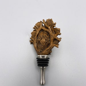 Op de afbeelding: Een houten flessenstop in de vorm van een hart met takken en bladeren. De stop is gemaakt van bruin hout en heeft een zilveren metalen basis.