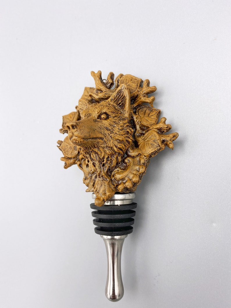 Op de afbeelding: Een bruine flesstopper van hars met een wolfskopontwerp omringd door bladeren en geweien. De stopper heeft een zilveren metalen basis.