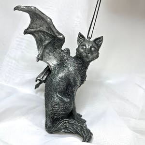May include: A silver-colored cat figurine with bat wings. The cat is sitting with its head turned to the side and its eyes are open. The figurine is hanging from a black cord.
