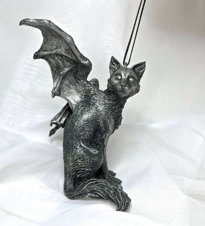 May include: A silver-colored cat figurine with bat wings. The cat is sitting with its head turned to the side and its eyes are open. The figurine is hanging from a black cord.