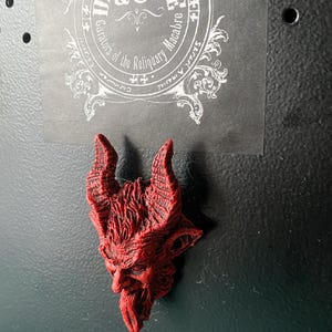 May include: Red devil head figurine with horns and a beard. The figurine is made of a textured material and has a detailed design. The figurine is attached to a black surface.