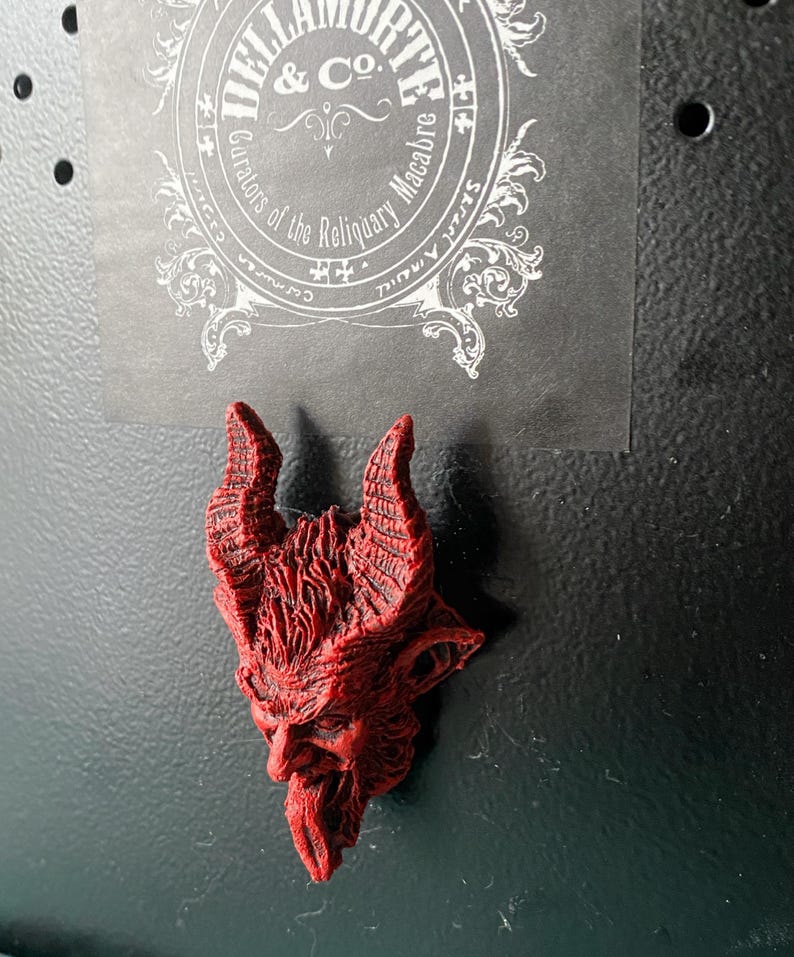 May include: Red devil head figurine with horns and a beard. The figurine is made of a textured material and has a detailed design. The figurine is attached to a black surface.