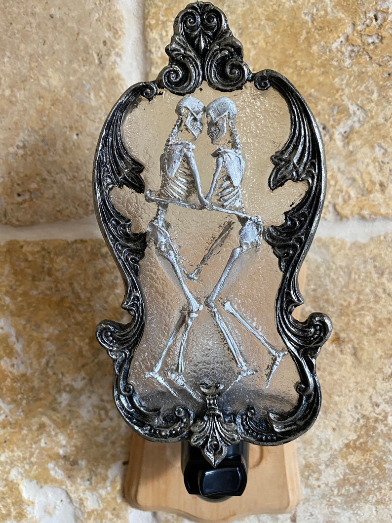 May include: A silver and black nightlight with a glass insert featuring two skeletons dancing. The skeletons are white and the glass is frosted. The nightlight is mounted on a wooden base.