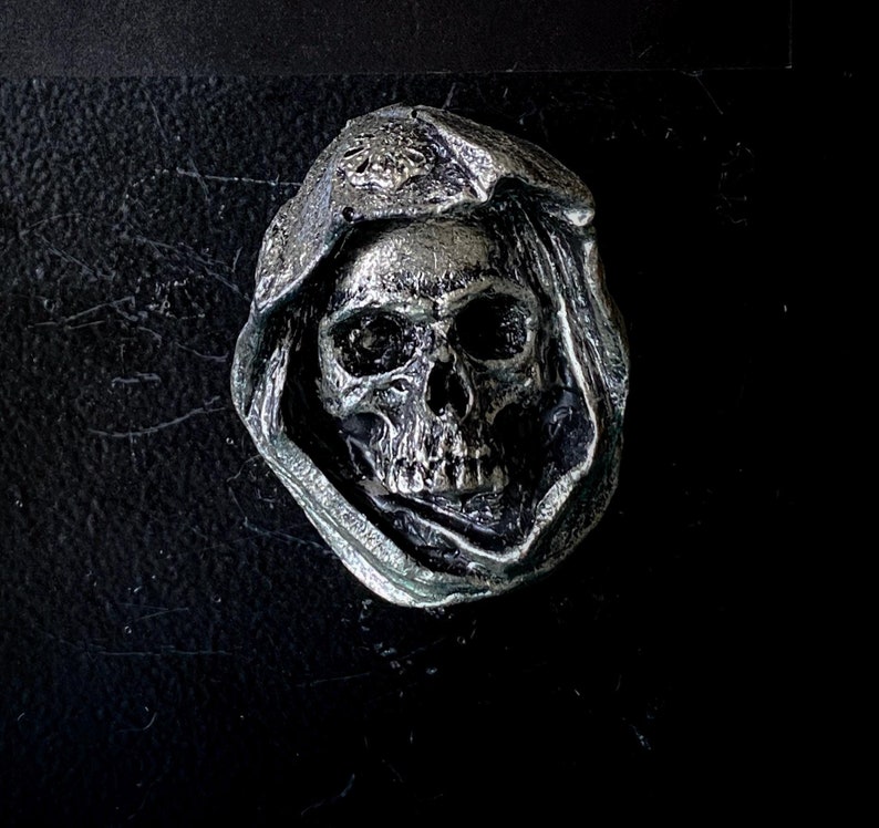 May include: A silver skull with a hooded cloak, the skull has a detailed design and is a decorative piece.