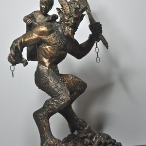 May include: A bronze sculpture of a goat-like creature with horns and a human-like body carrying a small child on its back. The creature is holding a chain and a staff in its hands. The sculpture is set on a rocky base.