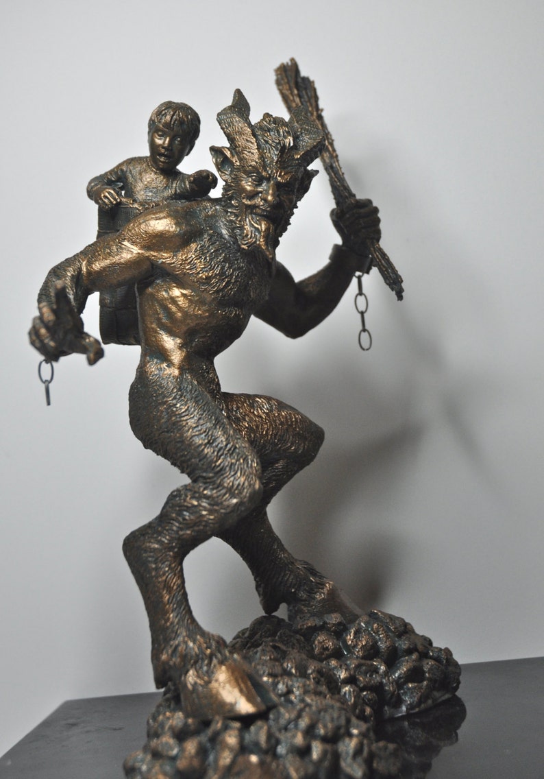 May include: A bronze sculpture of a goat-like creature with horns and a human-like body carrying a small child on its back. The creature is holding a chain and a staff in its hands. The sculpture is set on a rocky base.
