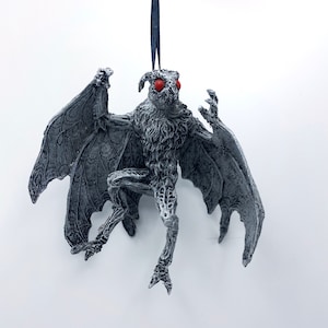 Puede incluir: Un adorno de pl&aacute;stico gris y blanco, detallado, de un mothman con ojos rojos. El mothman est&aacute; colgado de una cuerda azul.