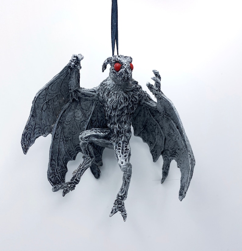 Puede incluir: Un adorno de pl&aacute;stico gris y blanco, detallado, de un mothman con ojos rojos. El mothman est&aacute; colgado de una cuerda azul.