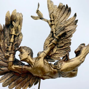 May include: A gold-toned sculpture of a winged figure with a muscular physique, flying with outstretched wings. The figure is attached to a base with a rough, textured surface.