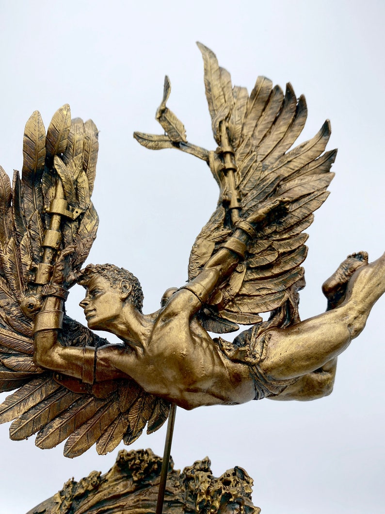 May include: A gold-toned sculpture of a winged figure with a muscular physique, flying with outstretched wings. The figure is attached to a base with a rough, textured surface.