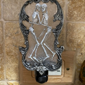 May include: A silver and black nightlight with a decorative frame and a clear glass insert featuring a skeletal couple dancing. The nightlight is plugged into an electrical outlet.