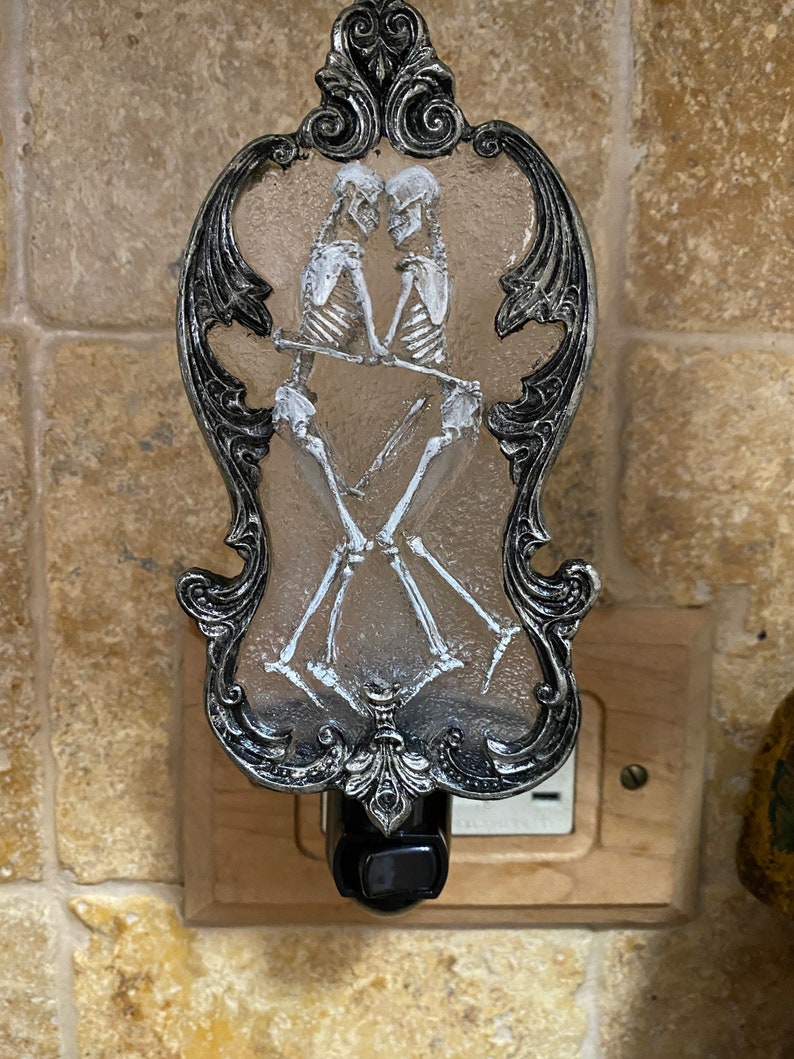 May include: A silver and black nightlight with a decorative frame and a clear glass insert featuring a skeletal couple dancing. The nightlight is plugged into an electrical outlet.