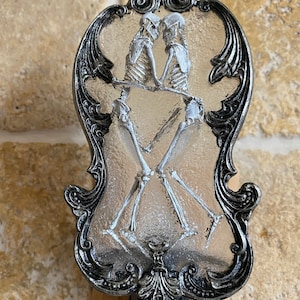 May include: A silver-coloured decorative night light with a clear glass insert featuring two skeletons holding hands. The skeletons are white and the glass is textured.