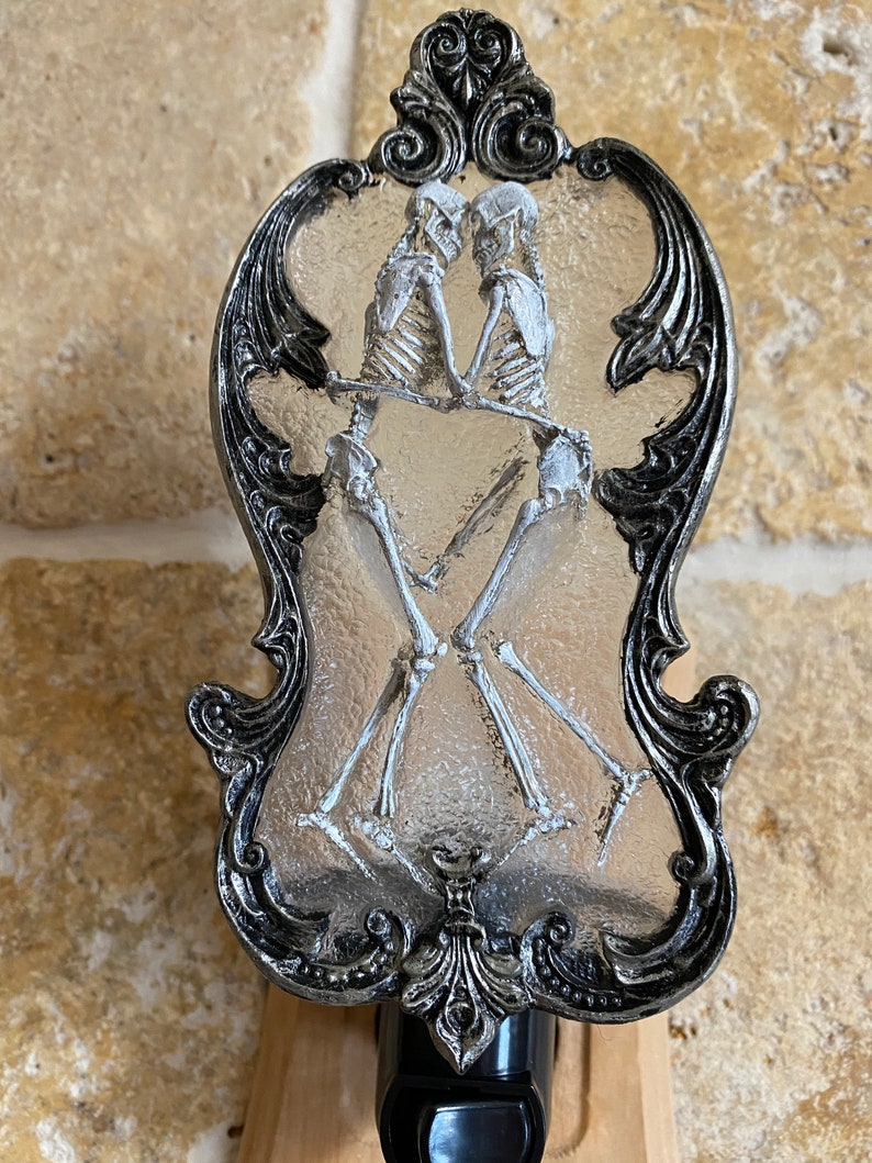 May include: A silver-coloured decorative night light with a clear glass insert featuring two skeletons holding hands. The skeletons are white and the glass is textured.