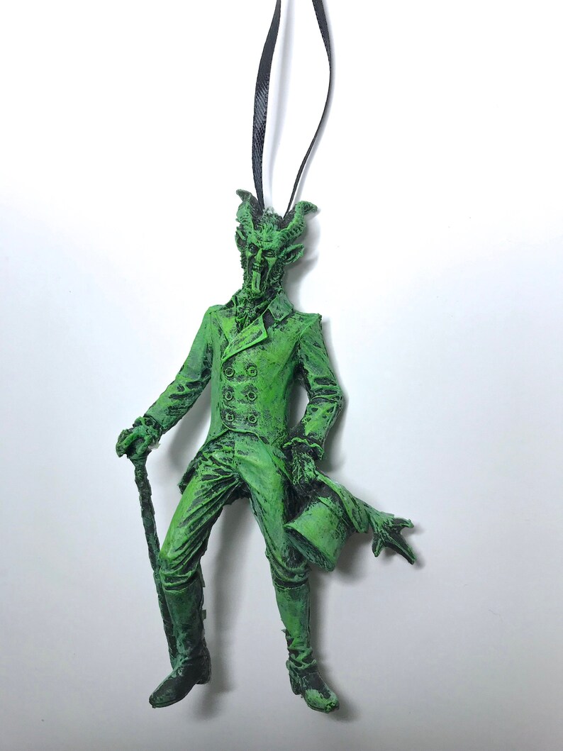 May include: Green Christmas ornament depicting a devilish gentleman in a top hat and coat, holding a cane. The ornament has a black string for hanging.
