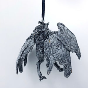 Puede incluir: Un adorno gris, detallado, con forma de murci&eacute;lago con un cord&oacute;n negro que cuelga de la parte superior. El murci&eacute;lago tiene un aspecto texturizado, casi esquel&eacute;tico, con intrincados dise&ntilde;os de alas.